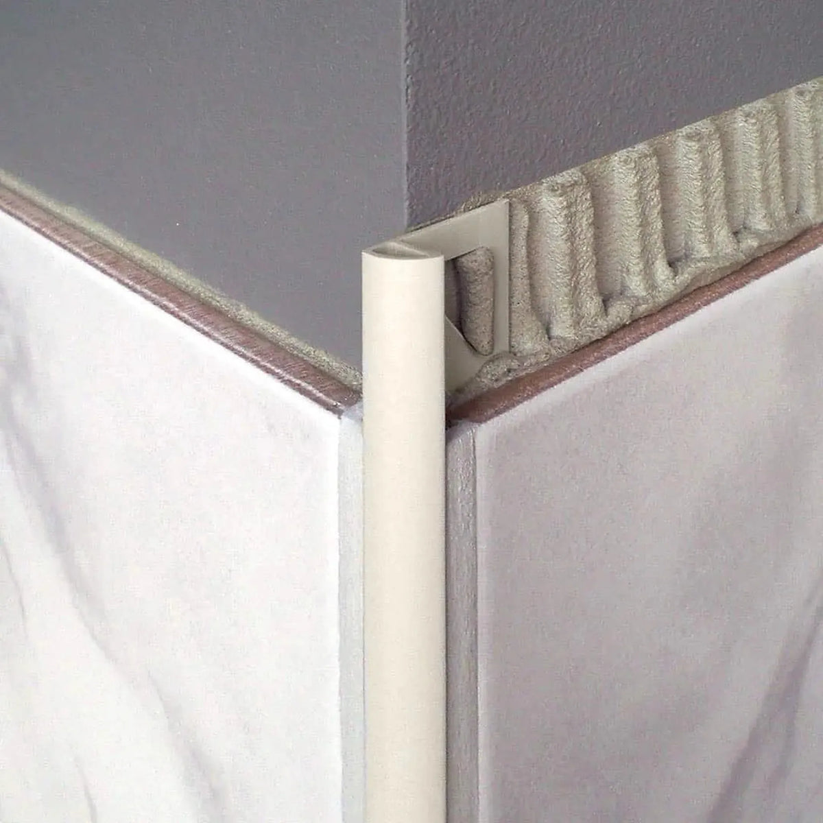 Profilé en quart de cercle en aluminium revêtu par poudrage couleur sable avec bord apparent arrondi, découpe triangulaire et aspect mat, posé à l'angle extérieur sous un carrelage clair.